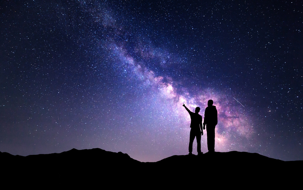 Milky Way with silhouette of a family on the mountain. Father and son pointing fingers in the night starry sky. Night landscape. Beautiful Universe. Space. Travel background with sky full of stars
