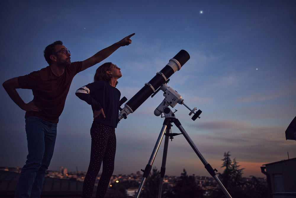 Silhouettes of father, daughter and astronomical telescope under starry skies