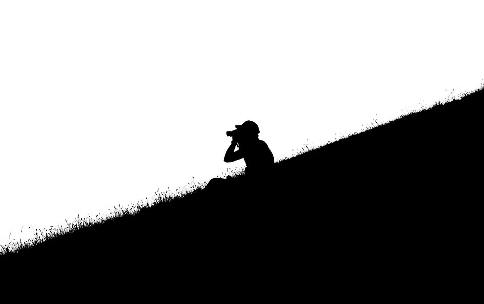 silhouette of a man sitting on a slope and holding a binocular and looking through it